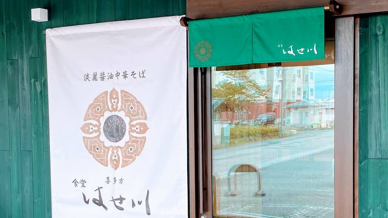 丸いロゴ入りの白いのれんと緑ののれんが掛かる食堂はせ川の入口